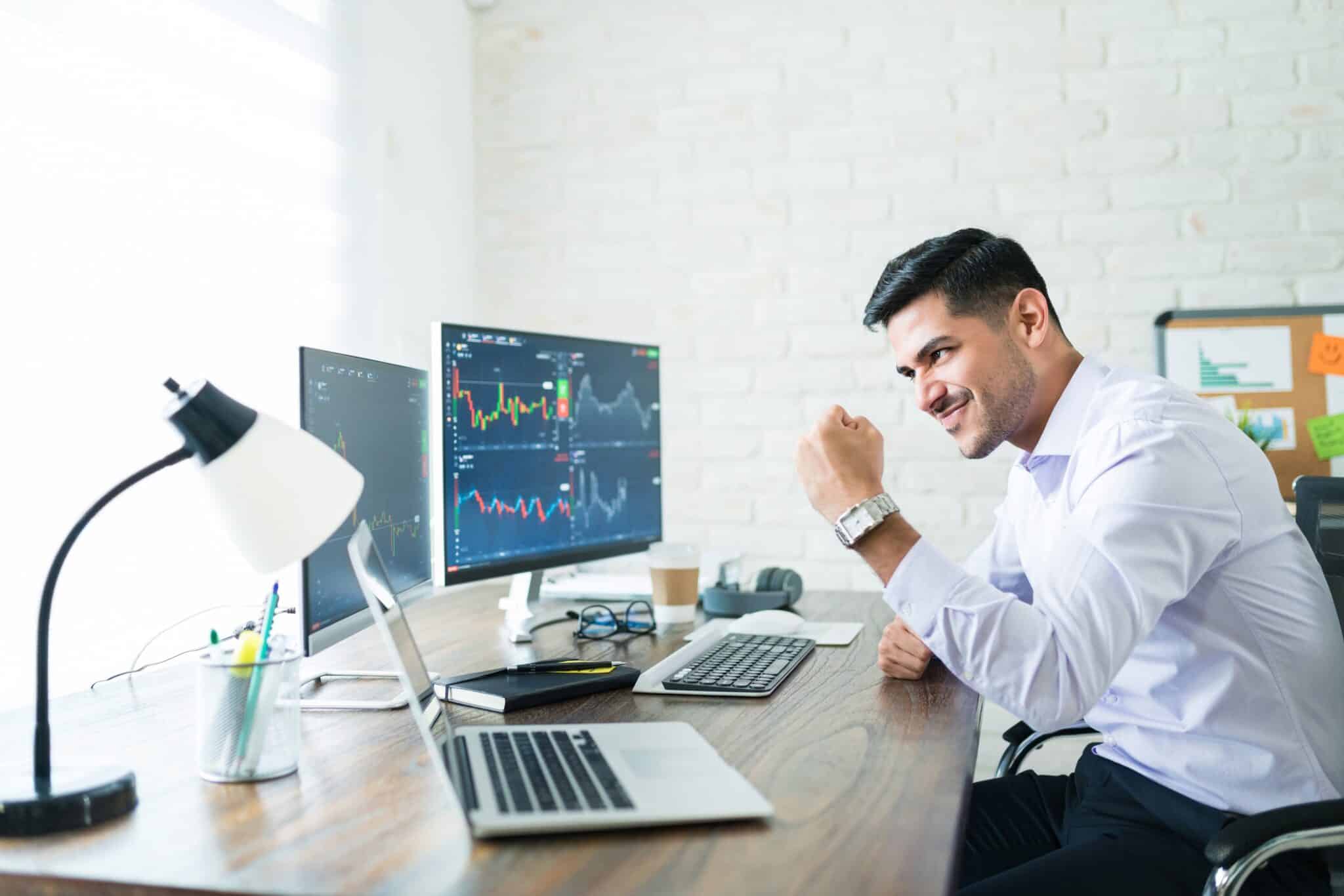 Confident handsome Hispanic financial broker showing fist to computer screen while trading from home